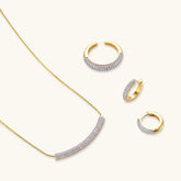 Gold-plated curved necklace, hoop earrings, and adjustable ring with pavé cubic zirconia stones on cream background.